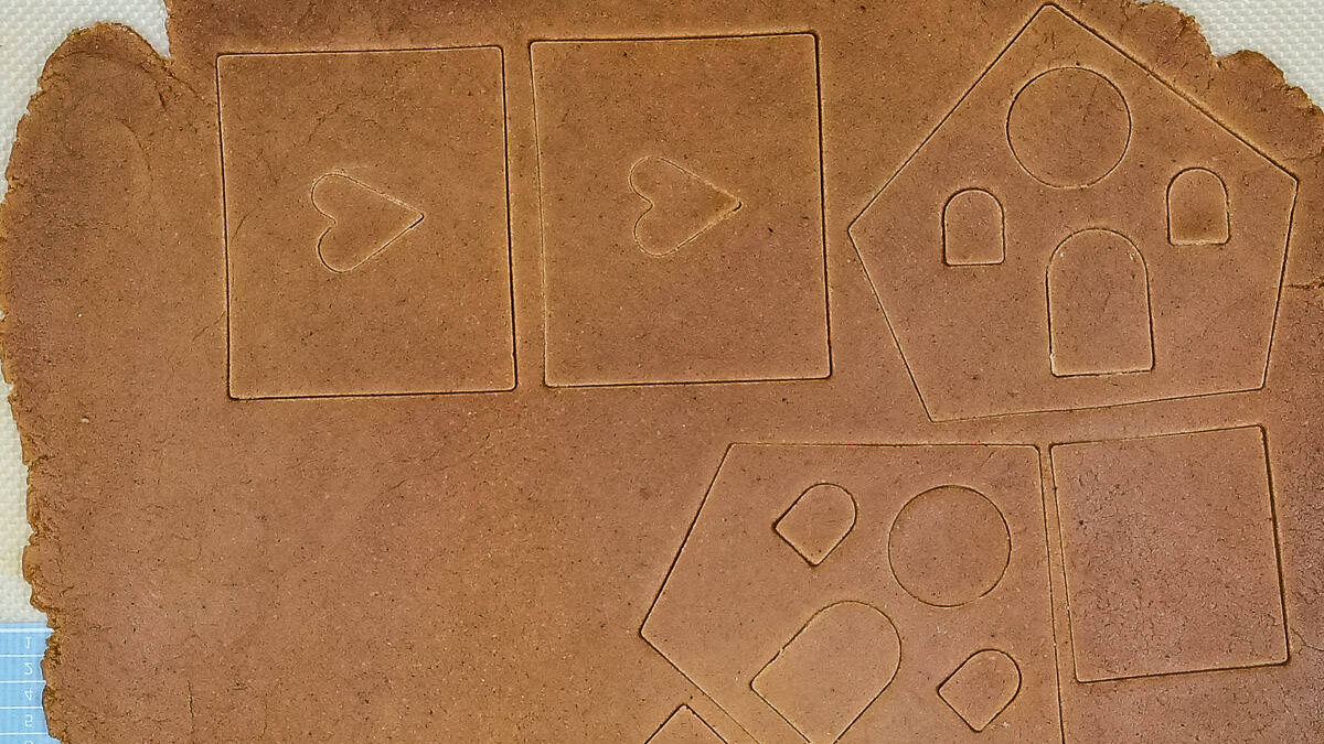 Rolled-out gingerbread dough with shapes for a Gingerbread House and rectangles with heart cutouts, ready to be cut and baked. The dough, perfect for your favorite gingerbread house recipe, rests on a flat surface with a textured background.