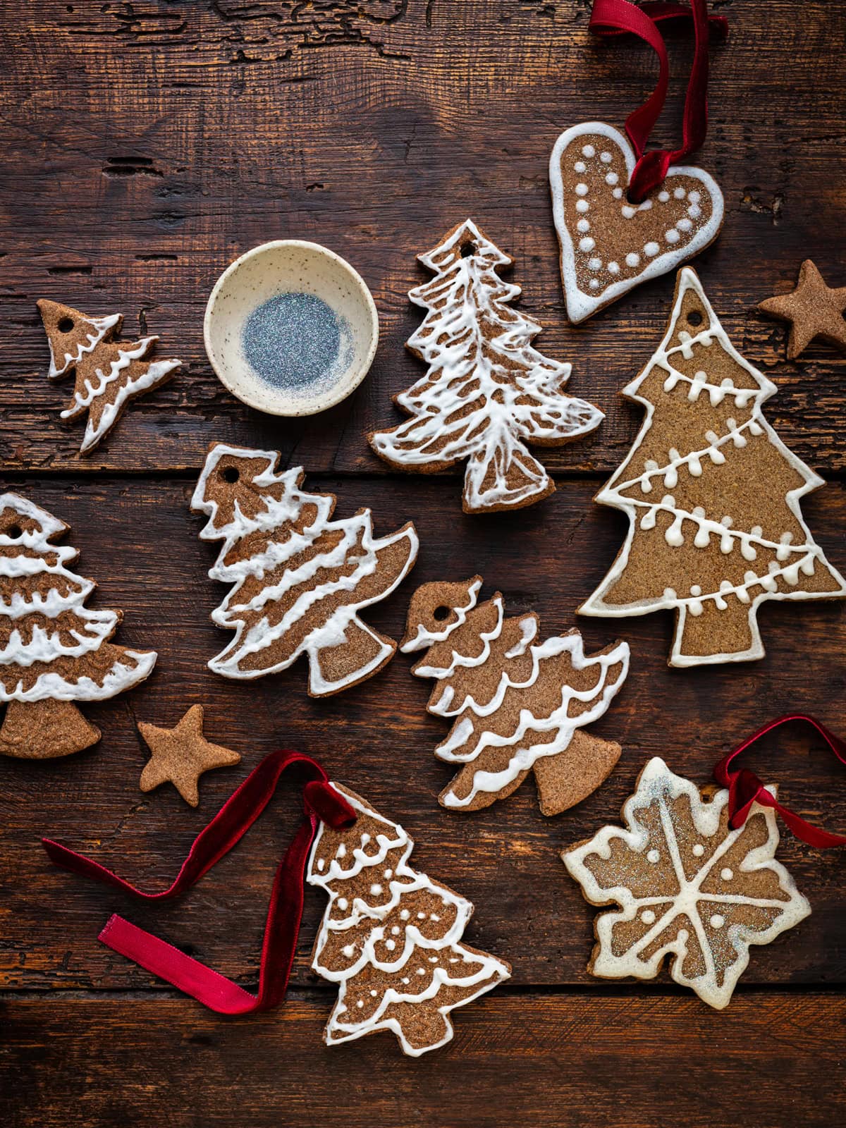 Decorated DIY gingerbread ornaments shaped like Christmas trees, stars, a heart, and a snowflake are arranged on a rustic wooden table, some with red ribbons and a small bowl of sugar nearby.