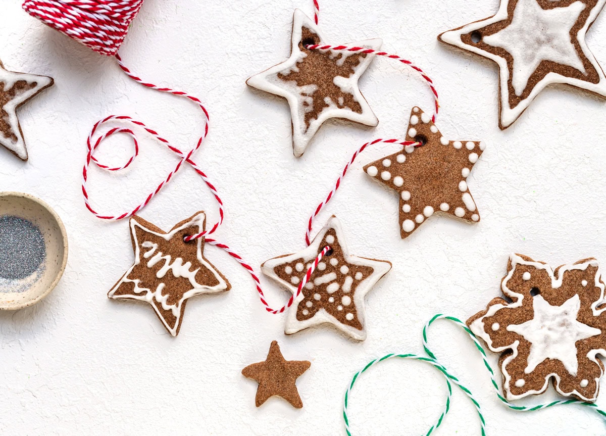 Star-shaped gingerbread ornaments with white icing are arranged on a white surface. Red and green string is threaded through some cookies, suggesting they can be used as ornaments. A spool of twine and a small bowl are nearby.
