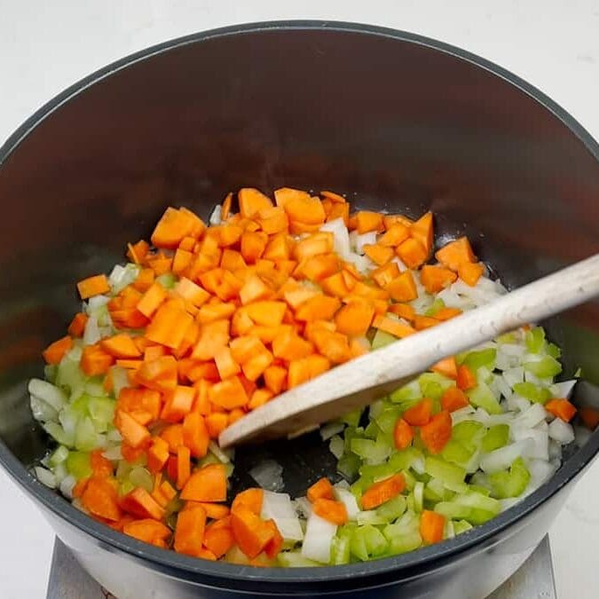 A pot on a stove contains chopped carrots, celery, and onions being stirred with a wooden spoon.