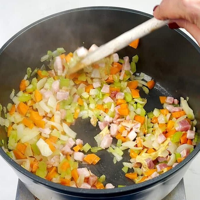 A hand stirs diced onions, carrots, celery, and bacon in a black pot with a wooden spoon as the ingredients sauté and steam rises.