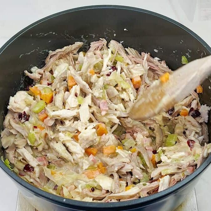 A pot filled with shredded chicken, diced carrots, celery, and cranberries being stirred with a wooden spoon. The mixture appears creamy, suggesting preparation for a savory dish.