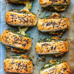 Eight golden Spinach Feta Puff Pastry Rolls filled with melted cheese and green pesto, topped with black sesame seeds, arranged on parchment paper. The flaky layers reveal some cheese deliciously oozing out.