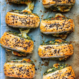 Eight golden Spinach Feta Puff Pastry Rolls filled with melted cheese and green pesto, topped with black sesame seeds, arranged on parchment paper. The flaky layers reveal some cheese deliciously oozing out.