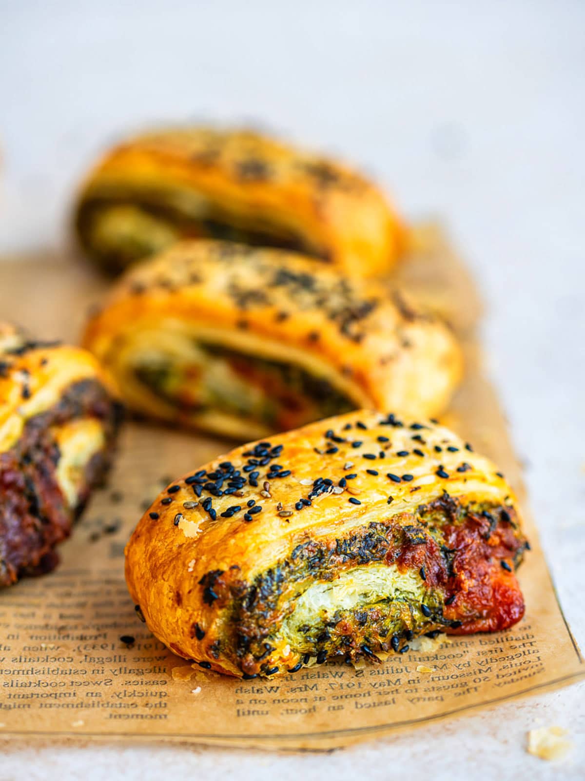 Golden Spinach Feta Puff Pastry Rolls filled with a green and red vegetable mixture, topped with black sesame seeds, rest on printed parchment paper. Three rolls are in the background, one in focus at the front.