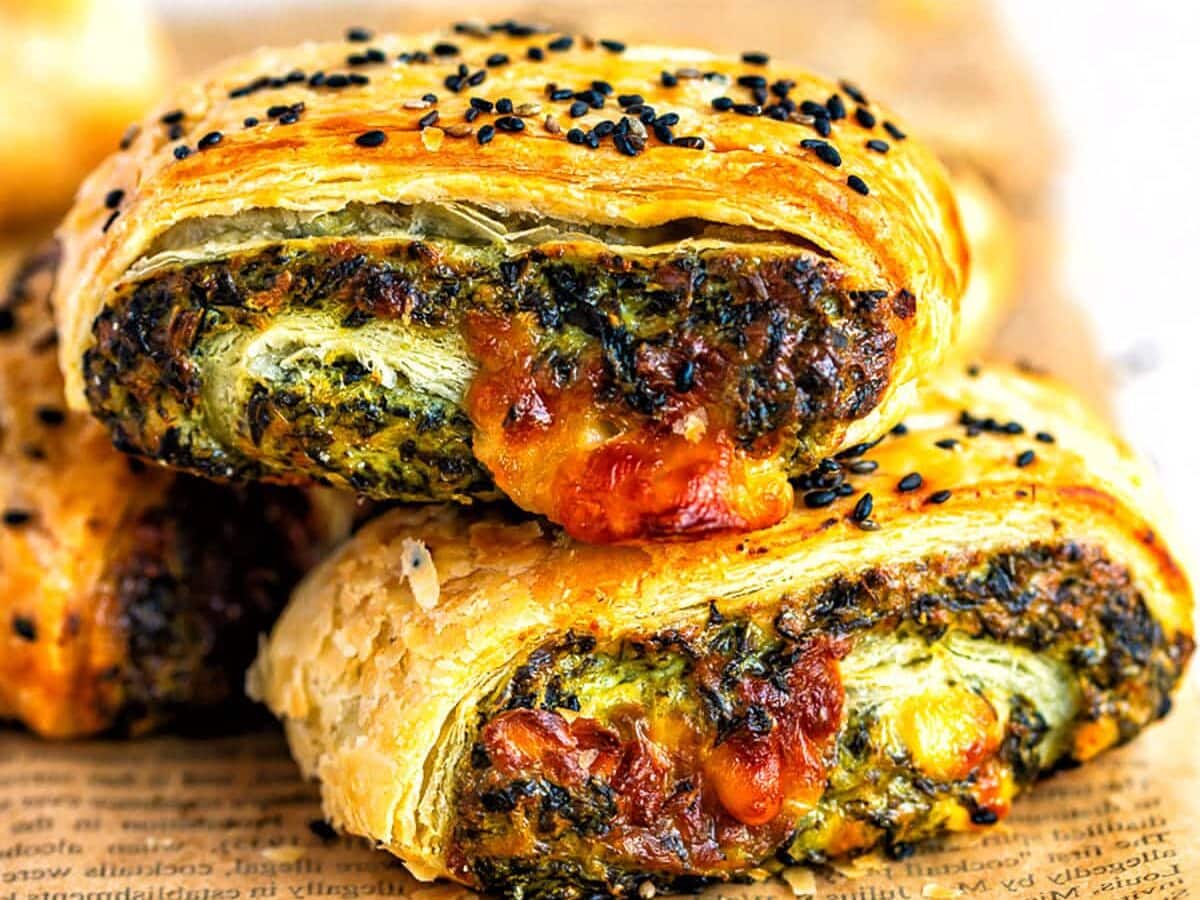 Two golden, flaky pastry rolls filled with spinach and feta cheese topped with black sesame seeds, are stacked on a piece of printed parchment paper.
