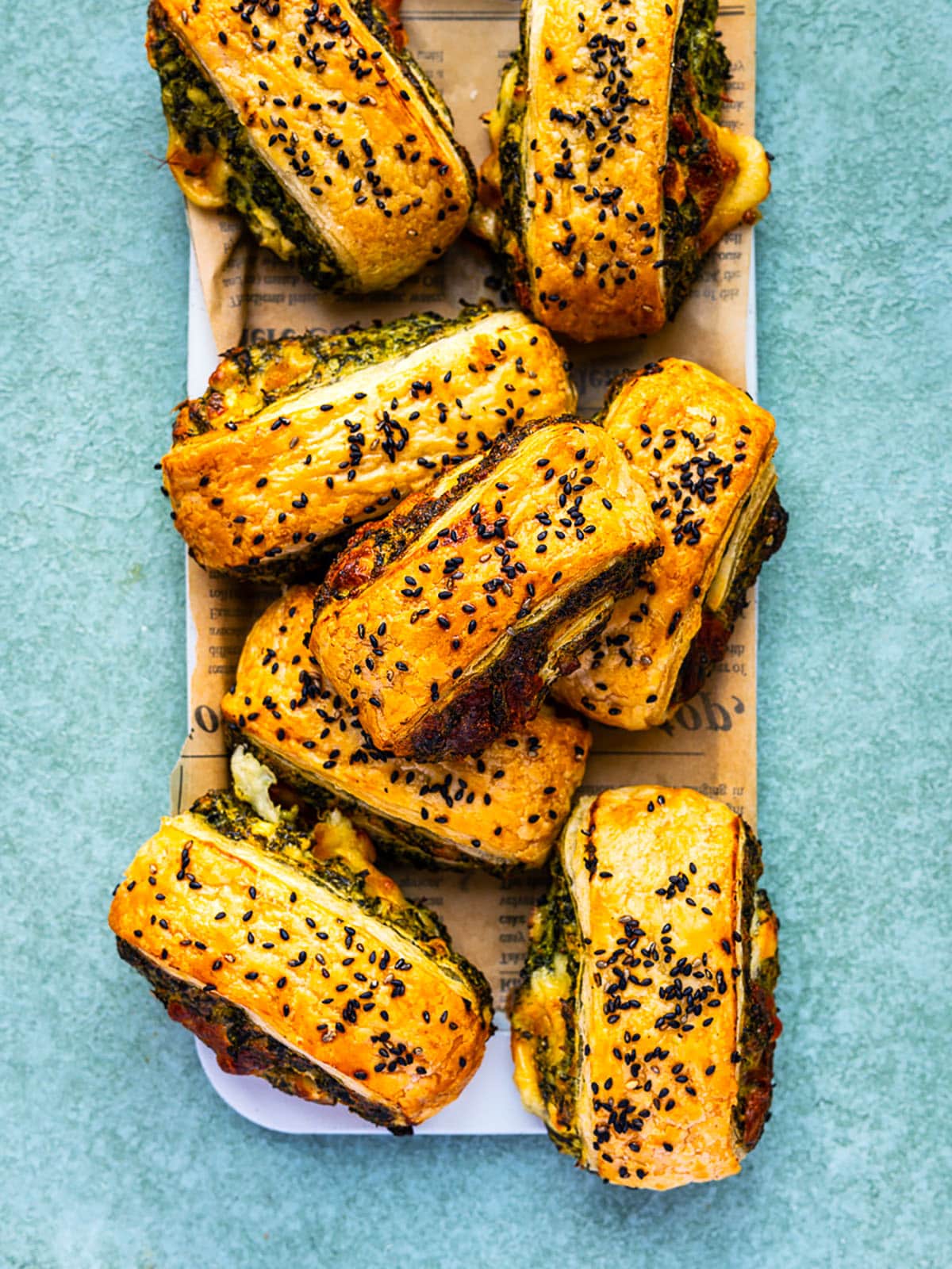 A platter of golden, flaky Spinach Feta Puff Pastry Rolls filled with green pesto and cheese, topped with black sesame seeds, displayed on parchment paper over a light blue surface.