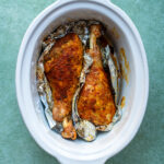 Two seasoned turkey legs wrapped in aluminum foil, placed inside a white oval slow cooker on a green surface.