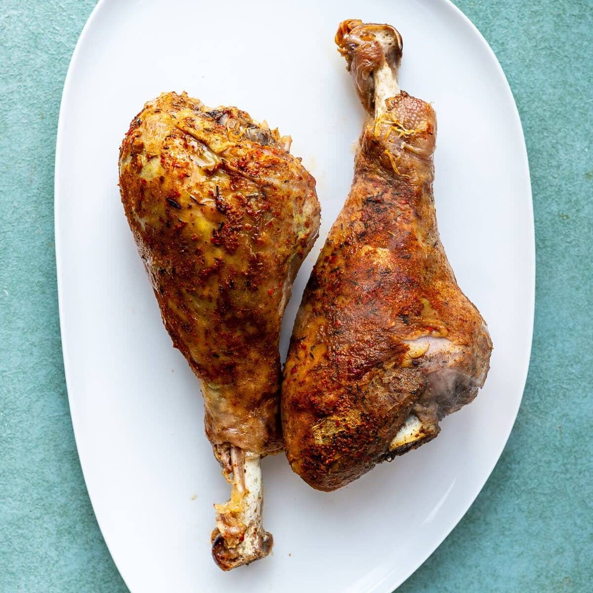 Two seasoned and roasted turkey legs placed side by side on a white oval plate, set against a light green background.
