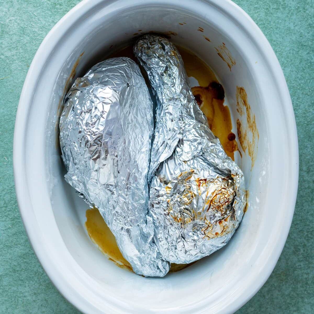 Two foil-wrapped items placed inside a white slow cooker with some brown cooking liquid and residue visible at the bottom. The slow cooker is set on a green surface.
