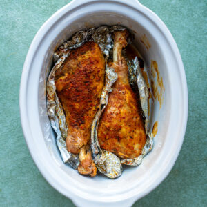 Two seasoned turkey legs wrapped in aluminum foil, placed inside a white oval slow cooker on a green surface.