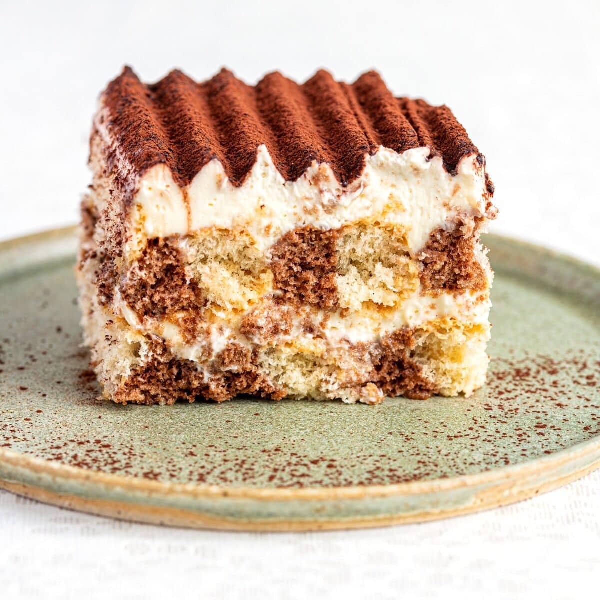 A slice of tiramisu sits on a green ceramic plate, showing layers of coffee-soaked sponge, creamy mascarpone, and a dusting of cocoa powder on top. The plate rests on a white lace-patterned tablecloth.