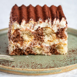 A slice of tiramisu with layers of creamy filling and coffee-soaked cake, topped with a dusting of cocoa powder, sits on a green ceramic plate next to a small fork.