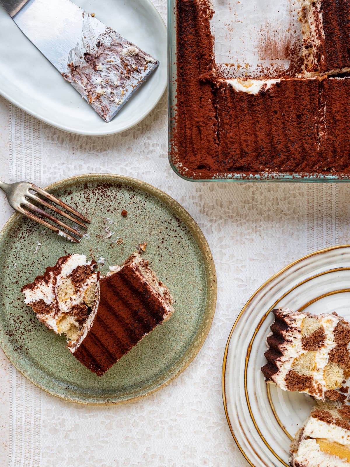 Top view of a partially sliced tiramisu in a glass dish, with two plated servings on green and white plates, a fork, and a cake server on a white lace tablecloth.