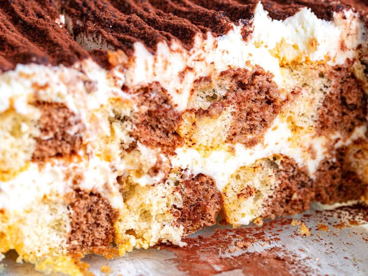 A close-up of a sliced tiramisu dessert in a glass dish, showing layers of coffee-soaked ladyfingers, creamy mascarpone filling, and cocoa powder dusted on top.