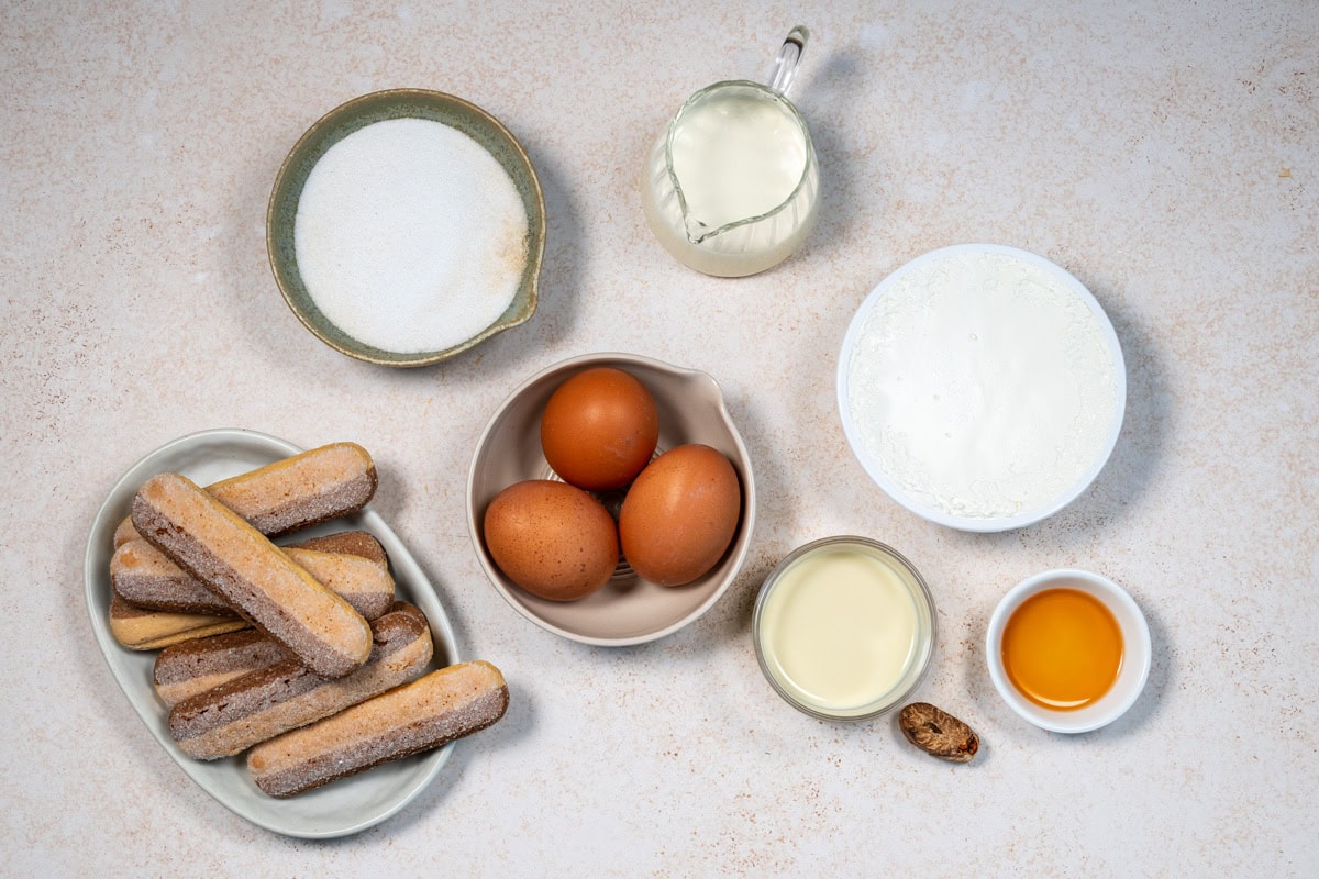 Ingredients for tiramisu are arranged on a light surface: sugar, milk, cream, three eggs, ladyfinger biscuits, sweetened condensed milk, a small dish of coffee liqueur, and a whole nutmeg.