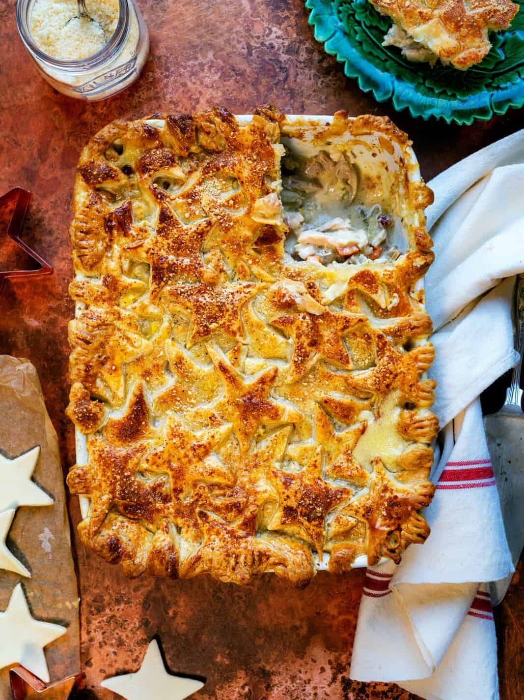 A golden-brown, rectangular pie topped with star-shaped pastry pieces sits on a brown surface. A slice has been removed, revealing a creamy filling, and a plate with a serving is nearby. Star-shaped pastry cutouts are beside the pie.