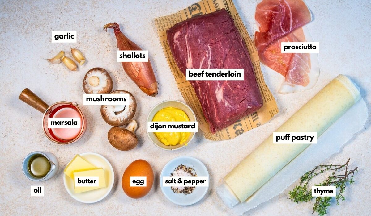 Top-down view of labeled ingredients for beef Wellington: garlic, shallots, mushrooms, marsala, oil, butter, egg, dijon mustard, salt & pepper, beef tenderloin, prosciutto, puff pastry, and thyme.