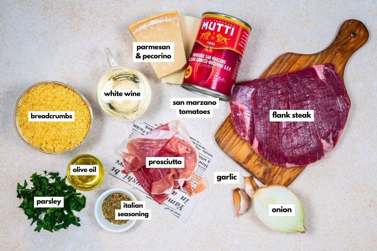 Ingredients for beef braciole recipe are laid out on a surface: flank steak, prosciutto, San Marzano tomatoes, parmesan & pecorino cheese, white wine, breadcrumbs, garlic, onion, parsley, olive oil, and Italian seasoning.