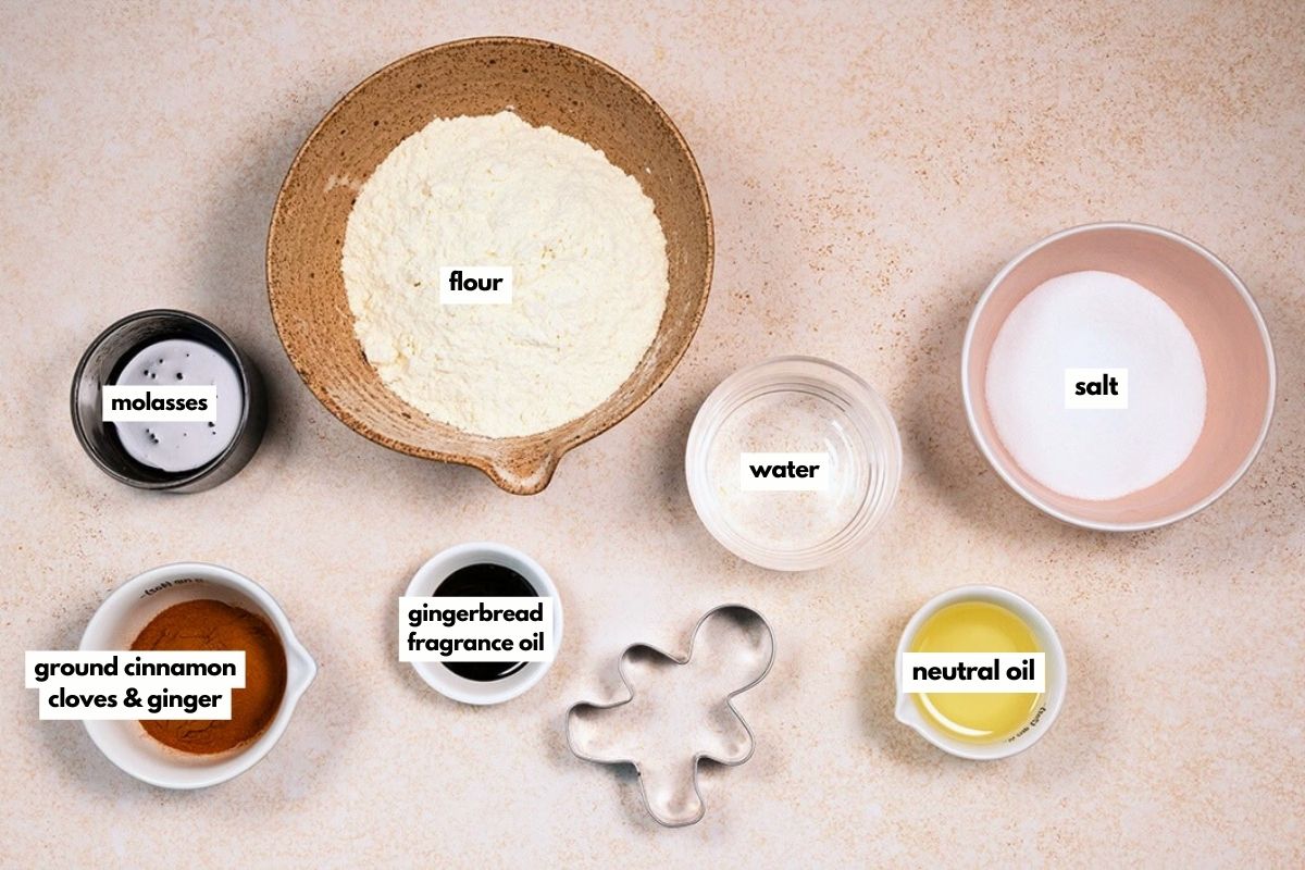 A top-down view of labeled ingredients for baking: flour, salt, water, molasses, ground cinnamon, cloves & ginger, gingerbread fragrance oil, neutral oil, and a gingerbread man-shaped cookie cutter.