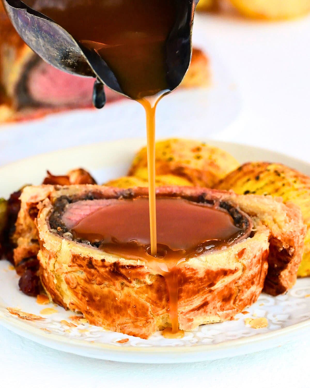 A close-up of a Beef Wellington slice on a plate, topped with rich brown sauce being poured over it, with roasted potatoes in the background—perfect inspiration if you're looking for the ultimate beef wellington recipe.