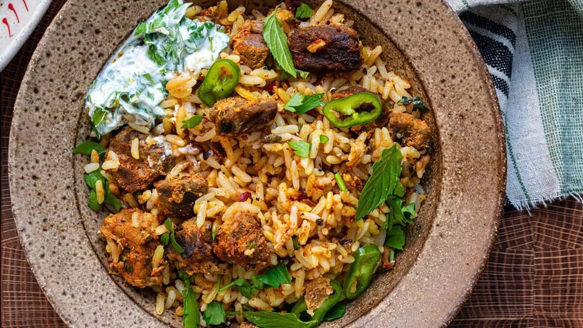A bowl of beef biryani with spiced rice, grilled meat, sliced green chili, and mint leaves, served alongside creamy herb yogurt sauce and a halved lime on a wooden surface. A striped cloth napkin and cutlery are nearby.