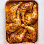 Baked chicken leg quarters with golden, crispy skin and herbs, arranged in a rectangular pan with a rich, seasoned sauce.