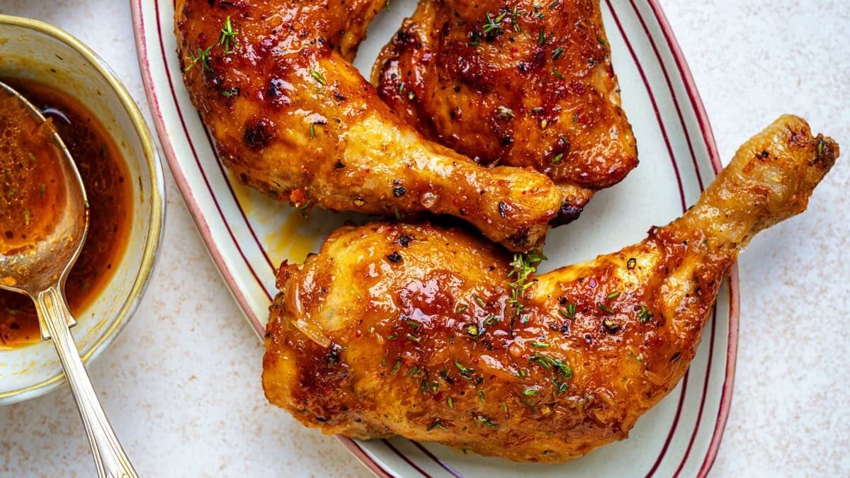 Three roasted chicken legs with golden, crispy skin are arranged on an oval plate. A bowl of sauce with a spoon and a small bowl of coarse salt are next to the plate on a light-colored surface.