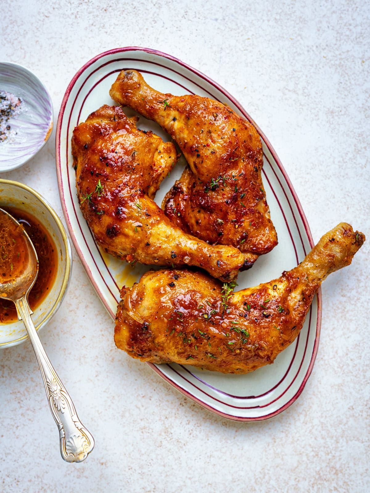 Three roasted chicken legs with golden, crispy skin are arranged on an oval plate. A bowl of sauce with a spoon and a small bowl of coarse salt are next to the plate on a light-colored surface.