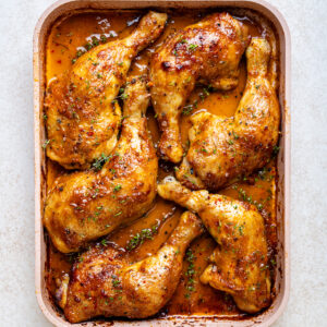 Baked chicken leg quarters with golden, crispy skin and herbs, arranged in a rectangular pan with a rich, seasoned sauce.