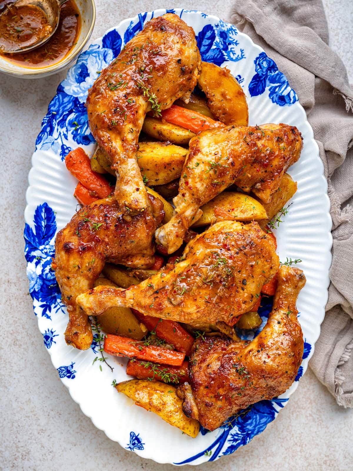 A platter with roasted chicken legs, golden potatoes, and carrots, garnished with herbs, served on a blue and white floral dish next to a beige napkin and a small bowl of sauce.