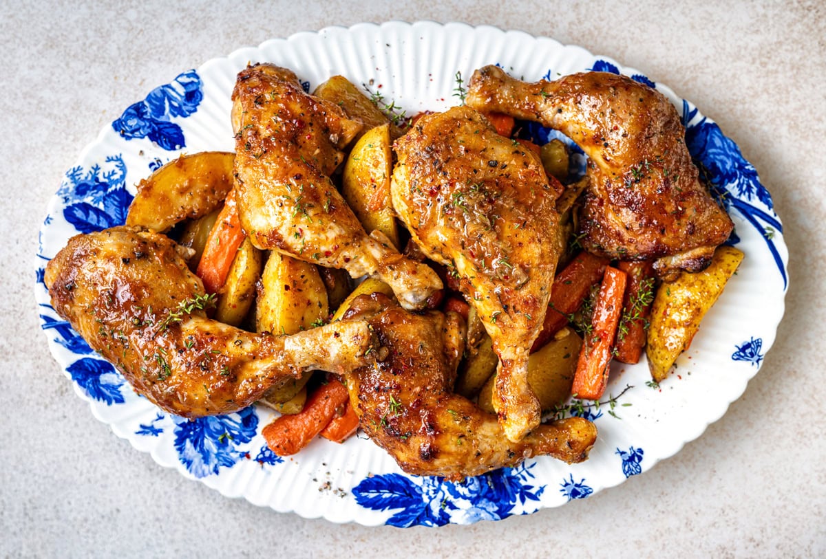 Four roasted whole chicken legs (chicken Marylands) served on a white plate with blue floral patterns, accompanied by roasted potato wedges and carrot pieces, garnished with herbs.
