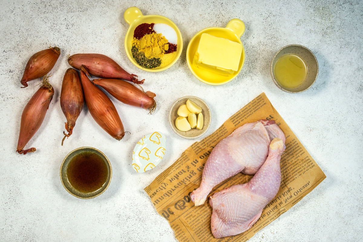Raw whole chicken legs on paper, shallots, bowls of spices, butter, garlic cloves, broth, and sauce arranged on a light surface.