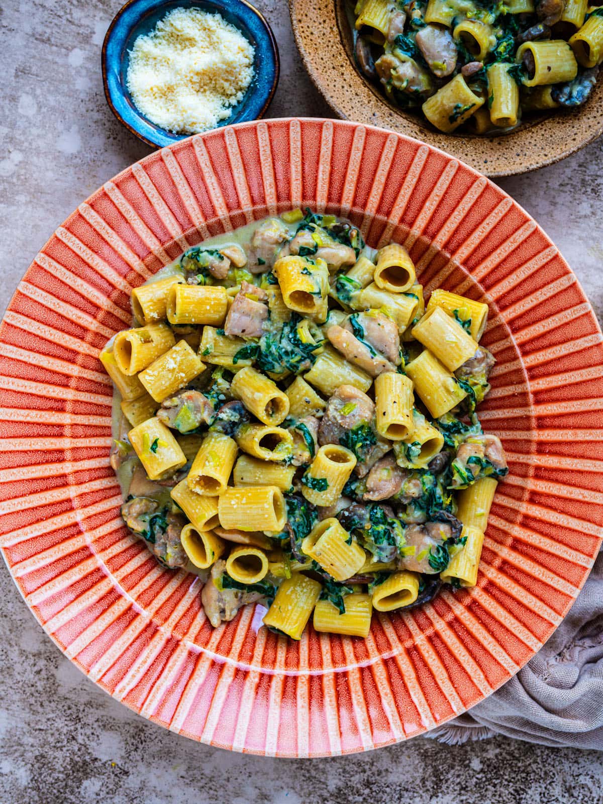 A red-rimmed plate filled with rigatoni pasta mixed with mushrooms, spinach, and a creamy sauce. A small bowl of grated cheese is nearby, and part of another bowl of pasta is visible in the corner.