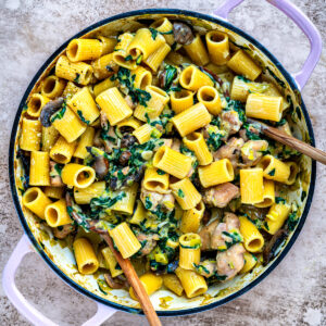 A large pot filled with rigatoni pasta, creamy sauce, mushrooms, diced chicken, fresh spinach, and parmesan. Two wooden spoons rest inside the pot on a textured surface.