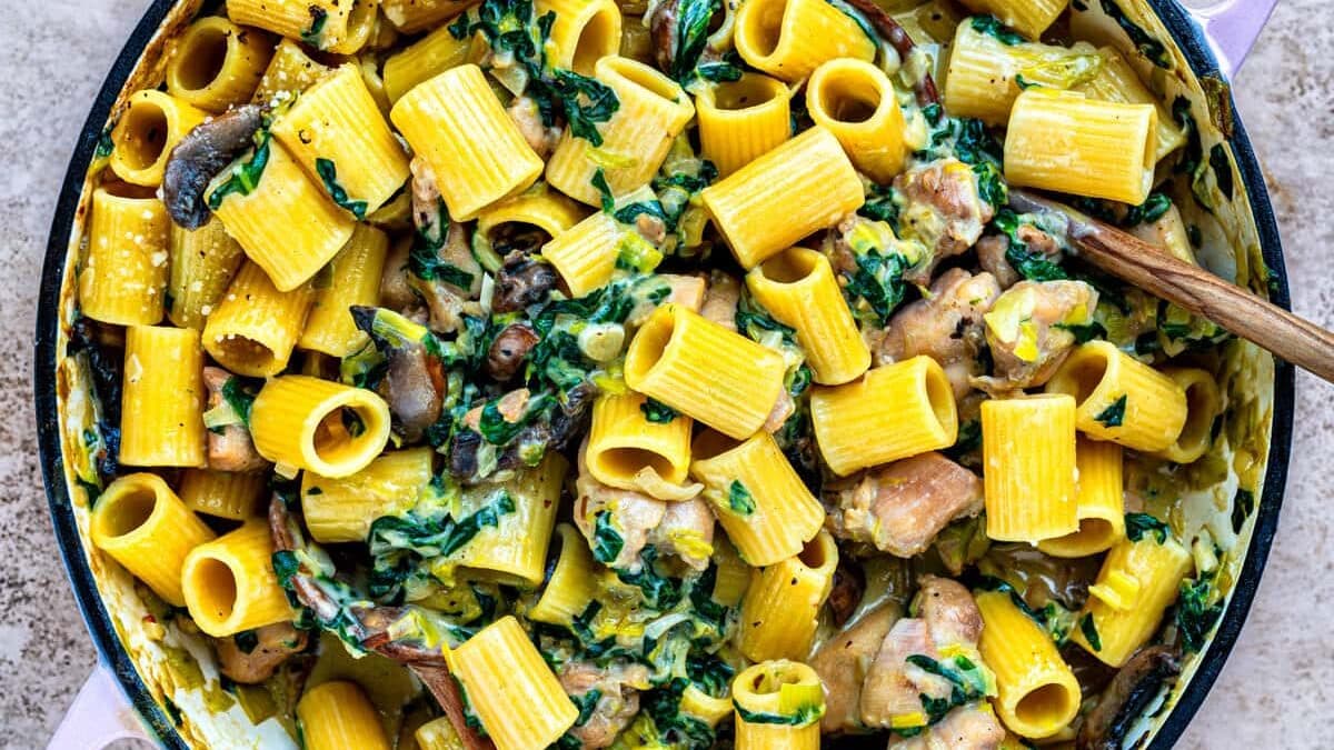 A large pot filled with rigatoni pasta, creamy sauce, mushrooms, diced chicken, fresh spinach, and parmesan. Two wooden spoons rest inside the pot on a textured surface.