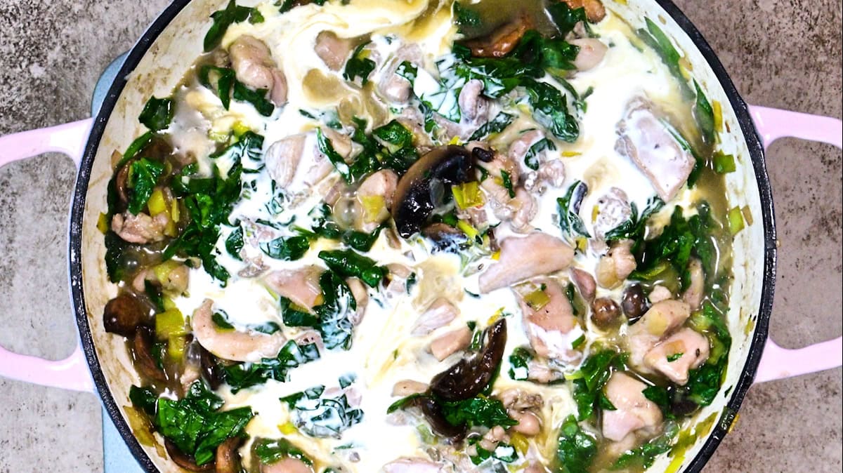 A creamy chicken and vegetable stew with spinach and mushrooms in a large pink-handled pot, viewed from above. The mixture features chunks of chicken, leafy greens, and slices of mushrooms in a white sauce.