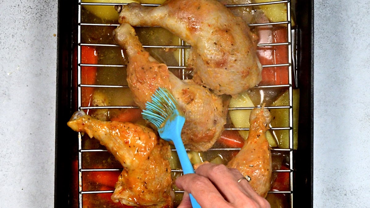 A hand uses a blue brush to glaze baked chicken legs on a wire rack, with roasted vegetables underneath in a baking tray.