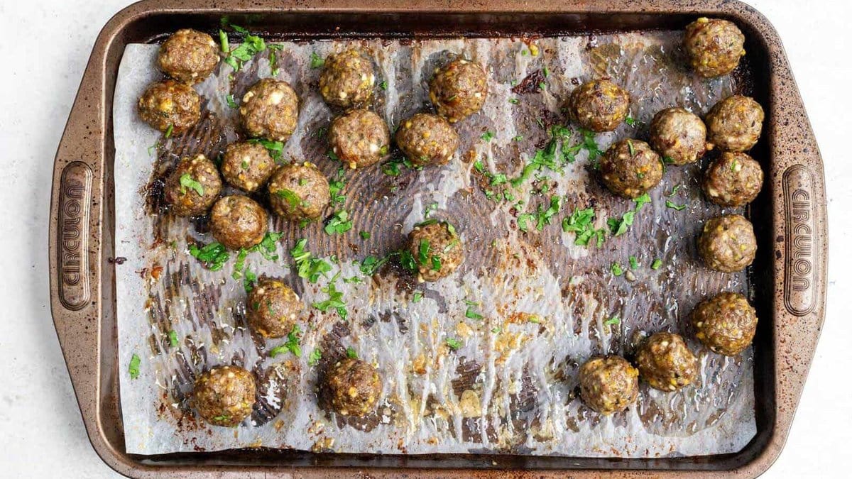 A baking tray lined with parchment paper holds about two dozen cooked meatballs, scattered irregularly, some garnished with chopped parsley. The tray shows signs of oil and browned spots from baking.