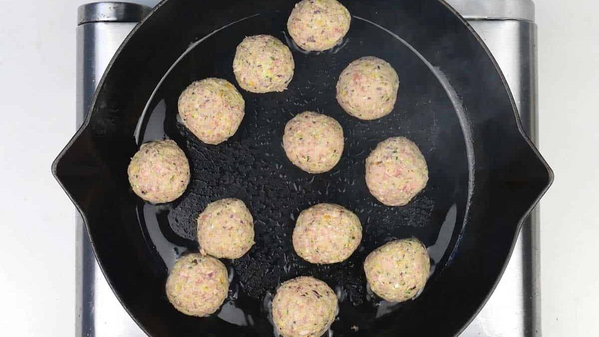 Raw meatballs are arranged in a circular pattern inside a black cast iron skillet, which is placed on a stovetop. The meatballs are light in color and appear to be uncooked.