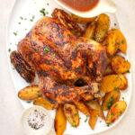 An oven roasted whole chicken garnished with herbs, served on a white platter with golden roasted potato wedges, a bowl of brown gravy, and a small dish of seasoned salt.