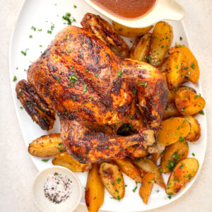 An oven roasted whole chicken garnished with herbs, served on a white platter with golden roasted potato wedges, a bowl of brown gravy, and a small dish of seasoned salt.