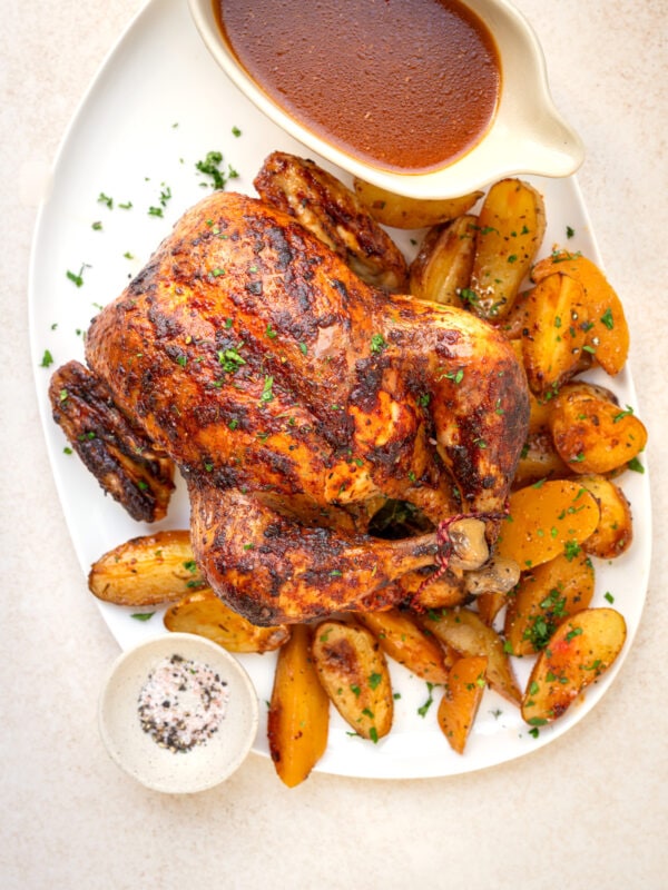 An oven roasted whole chicken garnished with herbs, served on a white platter with golden roasted potato wedges, a bowl of brown gravy, and a small dish of seasoned salt.