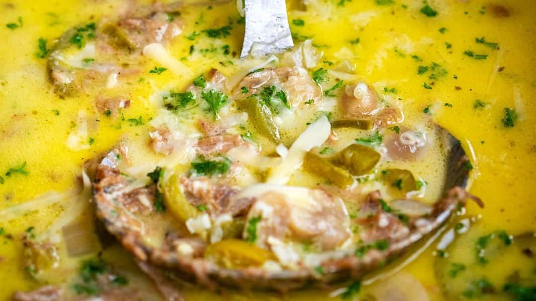 A close-up of a ladle lifting creamy, yellow-green Philly Cheesesteak Soup with ground meat, sliced pickles, and fresh herbs from a pot. The soup appears rich and hearty, with melted cheese and visible seasonings.