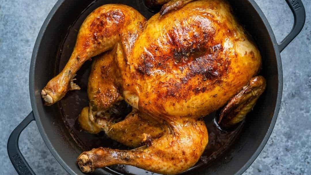 A Dutch oven roast chicken with golden brown, crispy skin sits in a black cast iron pot on a gray textured surface.