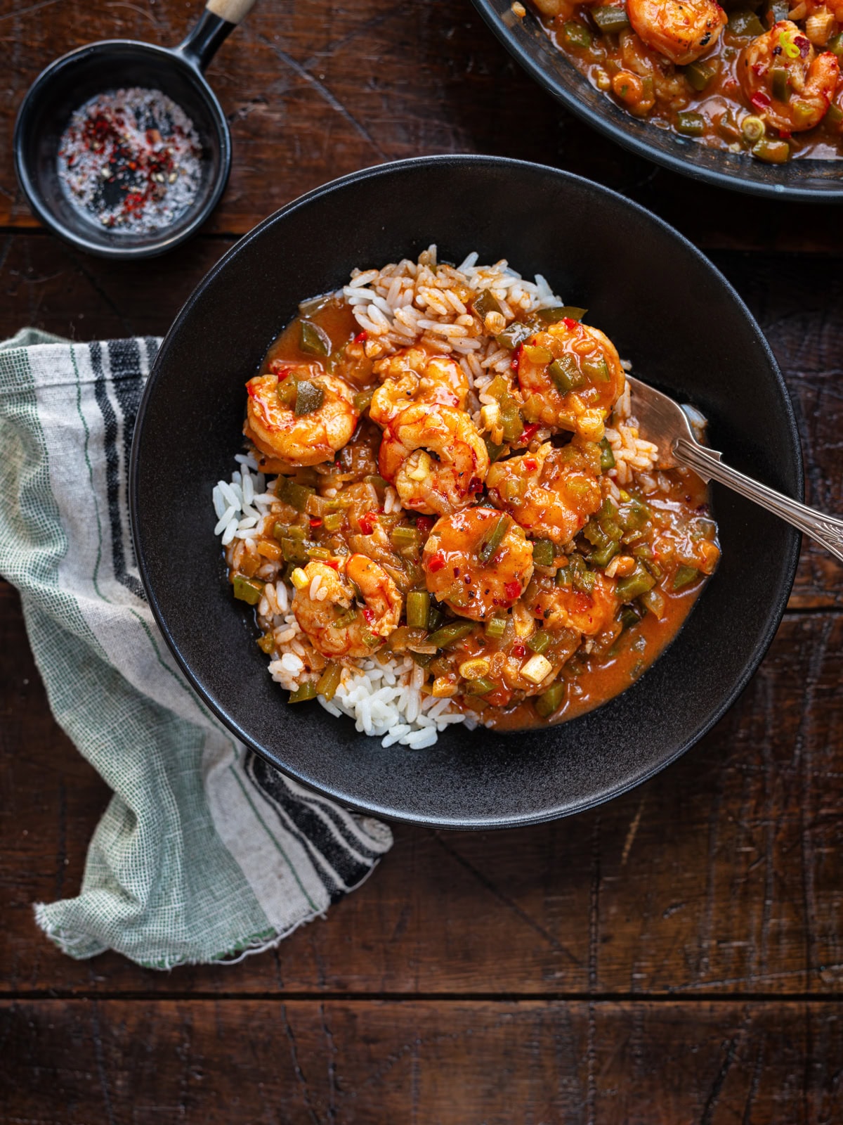 A black bowl filled with rice topped with shrimp and vegetable stew sits on a wooden table next to a striped cloth and a small black dish of seasoning. A spoon rests in the bowl.