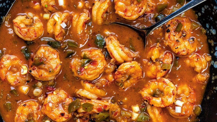 A bowl of Cajun shrimp stew in a rich, reddish sauce with herbs and vegetables, served on a rustic wooden table. A spoon rests in the bowl, with rice and spices nearby for an authentic Shrimp Étouffée recipe experience.