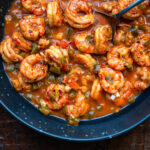 A close-up of a blue bowl filled with Cajun shrimp etouffee in a spicy, reddish-brown sauce with diced green peppers, and a spoon resting in the dish, all on a wooden surface.