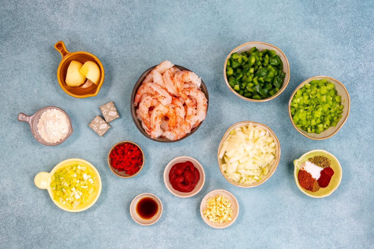 An overhead view of bowls and cups filled with chopped shrimp, green pepper, celery, onions, scallions, tomato paste, garlic, spices, stock cubes, flour, butter, and chopped red pepper on a blue surface.