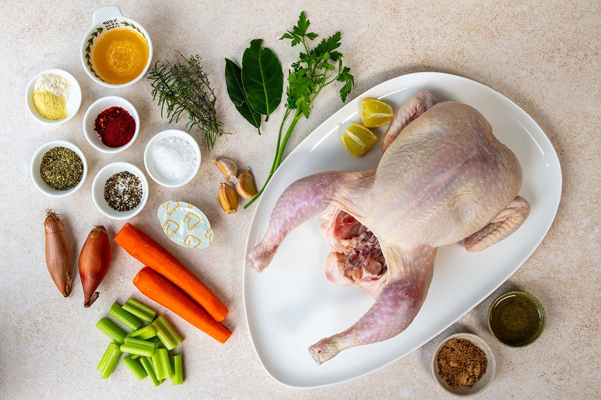 A whole raw chicken on a white platter with lemon wedges, surrounded by small bowls of spices, herbs, oil, salt, pepper, garlic, shallots, carrots, celery, and fresh parsley and bay leaves on a light surface.